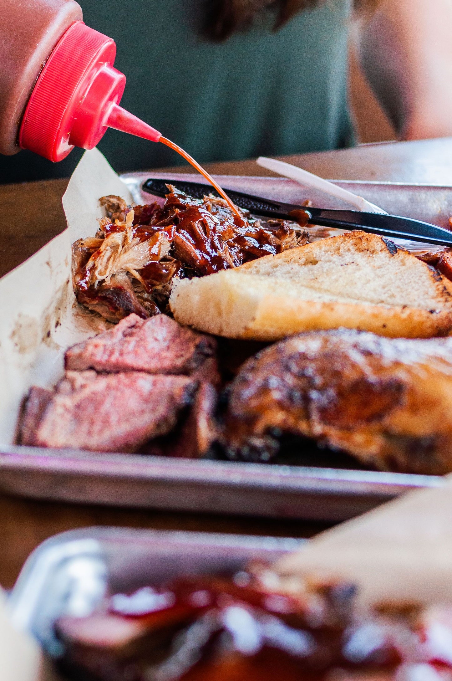 Spicy barbecue sauce being poured onto pulled pork alongside some garlic bread, tri tip, and chicken breasts.