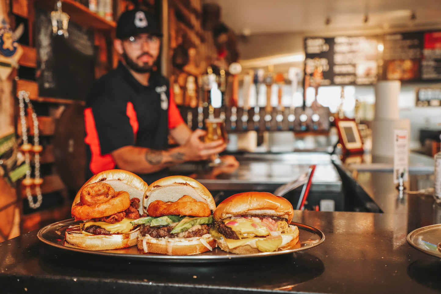 Three burgers with bacon, avocado, onion rings, cheese and pickles on a platter placed in front of the bartender who is holding a beer at Corbin's Q Bar and Barbecue. 