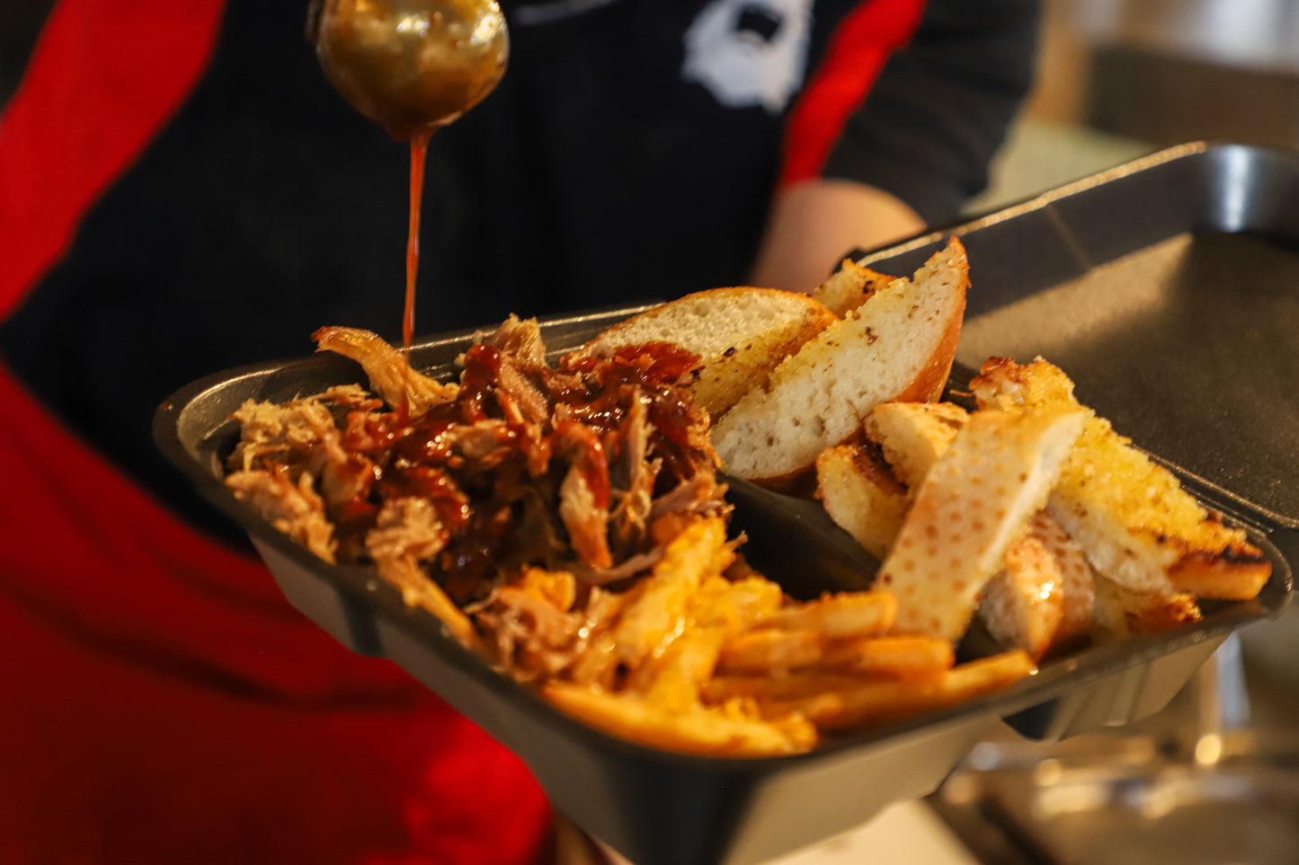 Sweet barbecue sauce being drizzled on some pulled pork in a to go box with garlic bread and french fries. 