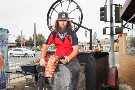 Corbin sitting on his Santa Maria style grill holding a full rack of pork ribs at his barbecue restaurant near SDSU.