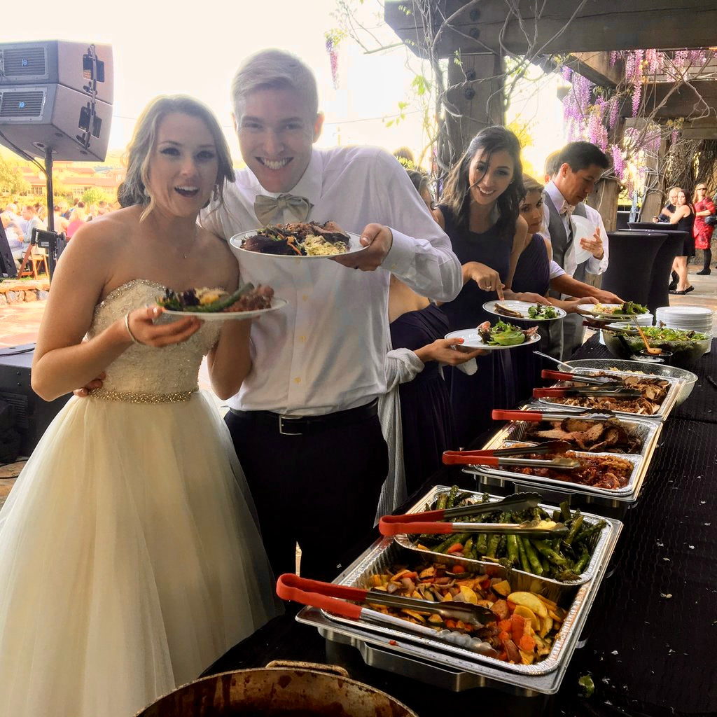 The bride and groom of a wedding hold up their plates after serving themselves from a buffet style on-site catering. 